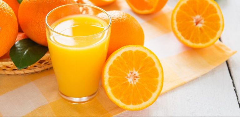 A glass of orange juice with sliced oranges around it on a table.