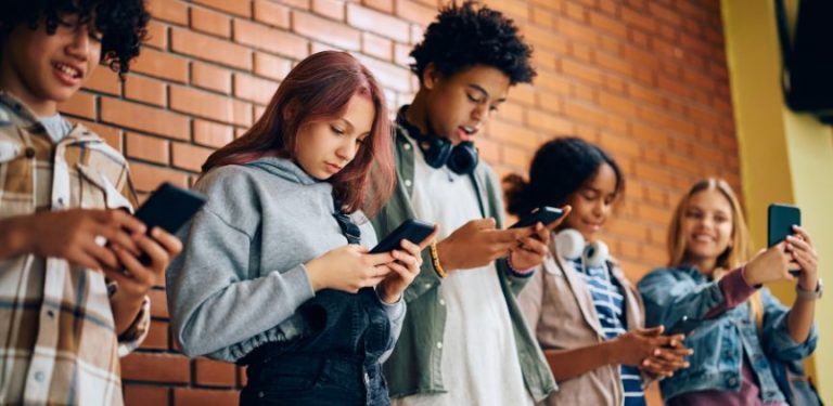 A group of teenagers standing against a wall, all using cell phones.