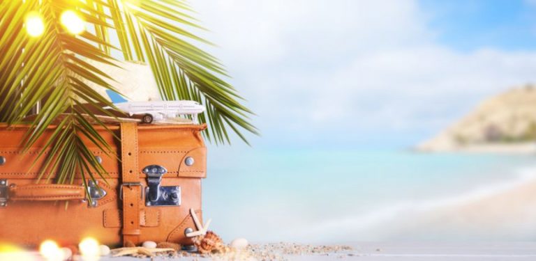 A brown suitcase on a beach, underneath palm tree leaves. There is a toy plane on top of it.