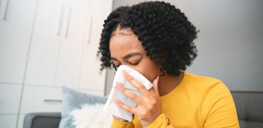 A Black woman in a yellow sweater blowing her nose in a tissue.