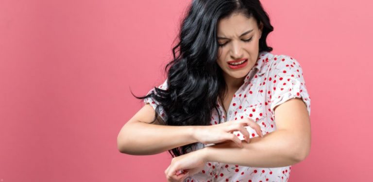 A woman wearing a white blouse with red cherries on it, standing in front of a pink background. She is looking down at her arm and scratching it.