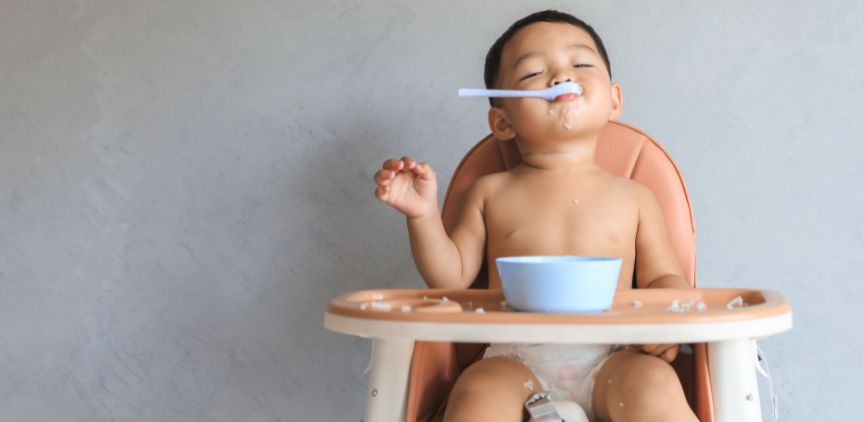 A baby in a highchair, eating food from a bowl with a spoon in their mouth.