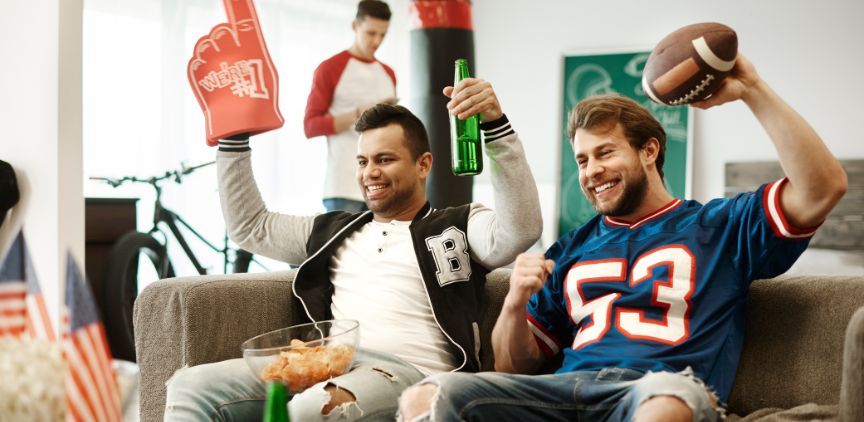 Two men sitting on a couch wearing football jerseys. They are cheering, holding a football and snacks, and watching a football game.